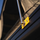 Close-up of a metal tent support pole attached to a yellow mounting bracket on a dark surface, holding up a tan fabric canopy—highlighting the strength and reliability of the CVT Mt. Hood Single Gas Strut.