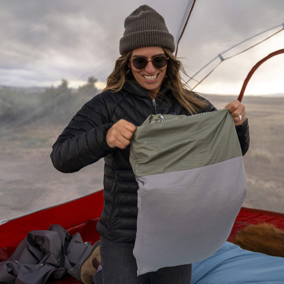 A smiling person in a beanie, sunglasses, and a puffer jacket stands in a tent, holding a gray and green camping bag. A sleeping pad and gear are on the tent floor. Cloudy sky and open landscape outside.