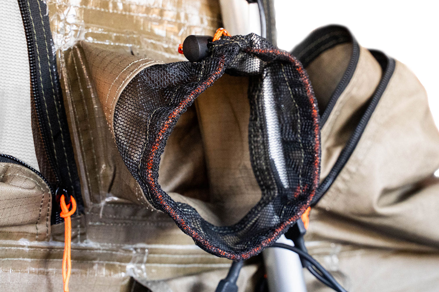 Close-up of a backpack pocket made of tan fabric with black mesh trim and orange stitching, featuring a drawstring closure and other visible compartments.