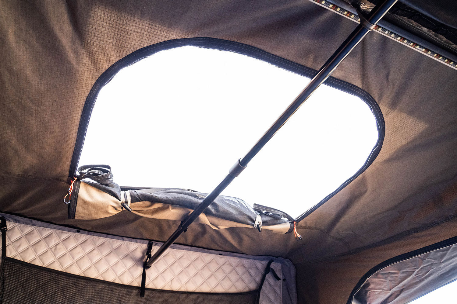 View from inside a rooftop tent looking up at an open skylight with a quilted interior lining, support poles, and a clear bright sky visible through the opening.