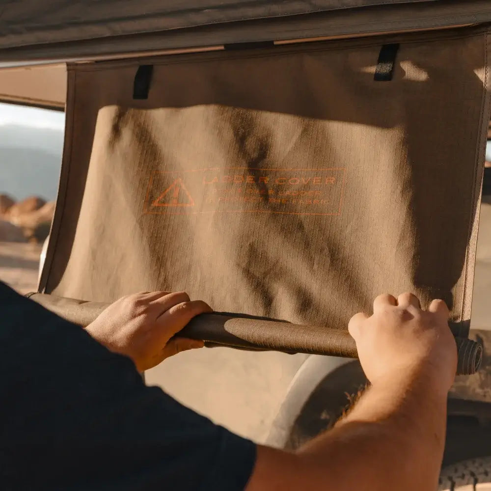 A person rolls up a tan fabric panel attached to a vehicle, outdoors. The panel, featuring a faint orange warning label, is part of the CVT Wasatch Hardshell Tent—an ideal rooftop tent setup for overland adventures or camping trips.