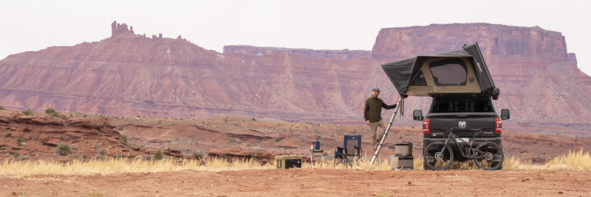 A person stands next to a black truck with a rooftop tent, parked on a desert plateau. Camping gear and a bike are nearby, with tall red rock mesas in the background.