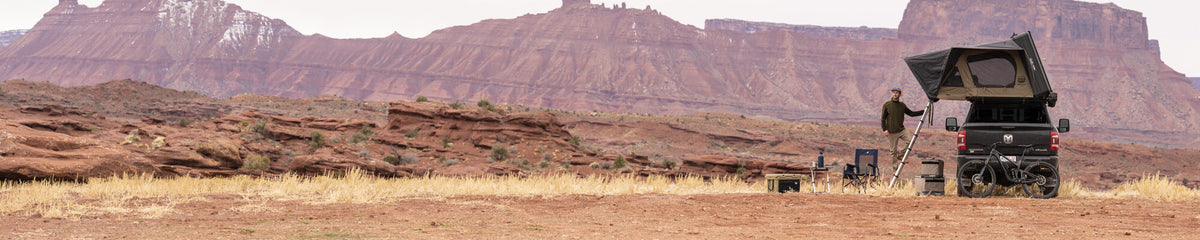 A person stands by a van with an open rooftop tent in a rocky desert landscape, surrounded by camping gear, with red cliffs and mesas in the background.