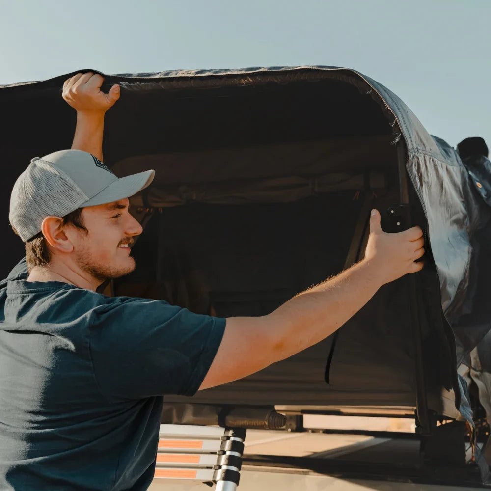 Wearing a gray cap and navy shirt, a person sets up the CVT Wasatch Hardshell Tent, unfolding its sturdy shell outdoors under a clear sky—preparing for their next overland adventure.