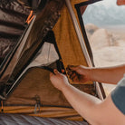 A person unzips a mesh window inside a CVT Wasatch Hardshell Tent, letting in light. The tent fabric is brown and the person’s hands are visible, adjusting the window flap—ready for another day of overland adventure.