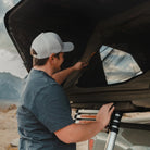 A man in a white cap and blue shirt sets up a CVT Wasatch Hardshell Tent on his vehicle, preparing for an overland adventure with mountains and cloudy skies in the background.