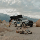 A person climbs a ladder to a CVT Wasatch Hardshell Tent mounted on a parked truck in a rocky desert landscape. Nearby are camping chairs, a fire ring surrounded by stones, and mountains rising in the background beneath a cloudy sky—creating the perfect setting for an unforgettable overland adventure.