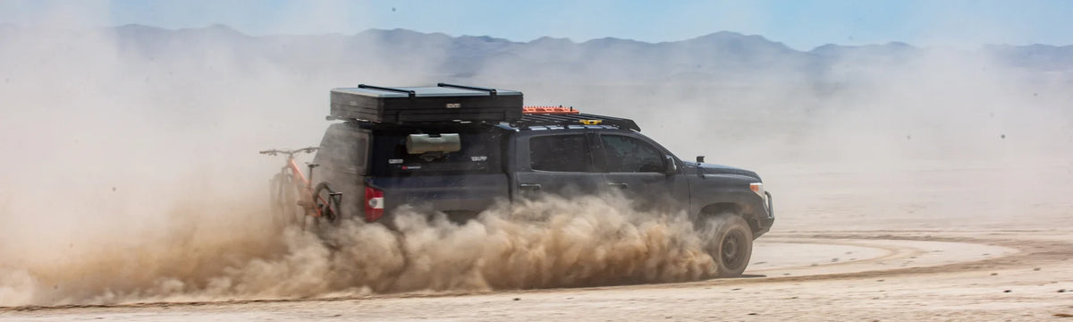 A black off-road truck with cargo and a bike drives fast on a dusty desert road, kicking up a large cloud of dust. Mountains are visible in the background under a hazy sky.