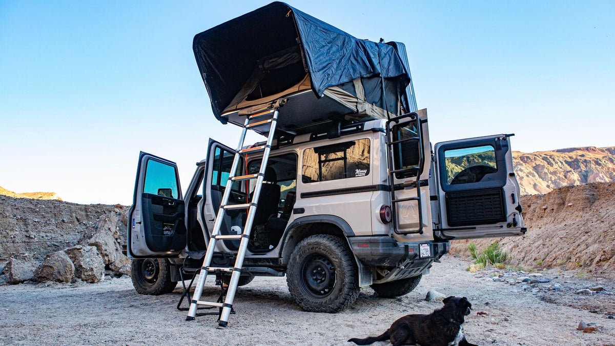 A white SUV with all doors open is parked on rocky ground. A rooftop tent with a ladder is extended. A black dog lies nearby. Rocky hills and a clear blue sky are in the background.