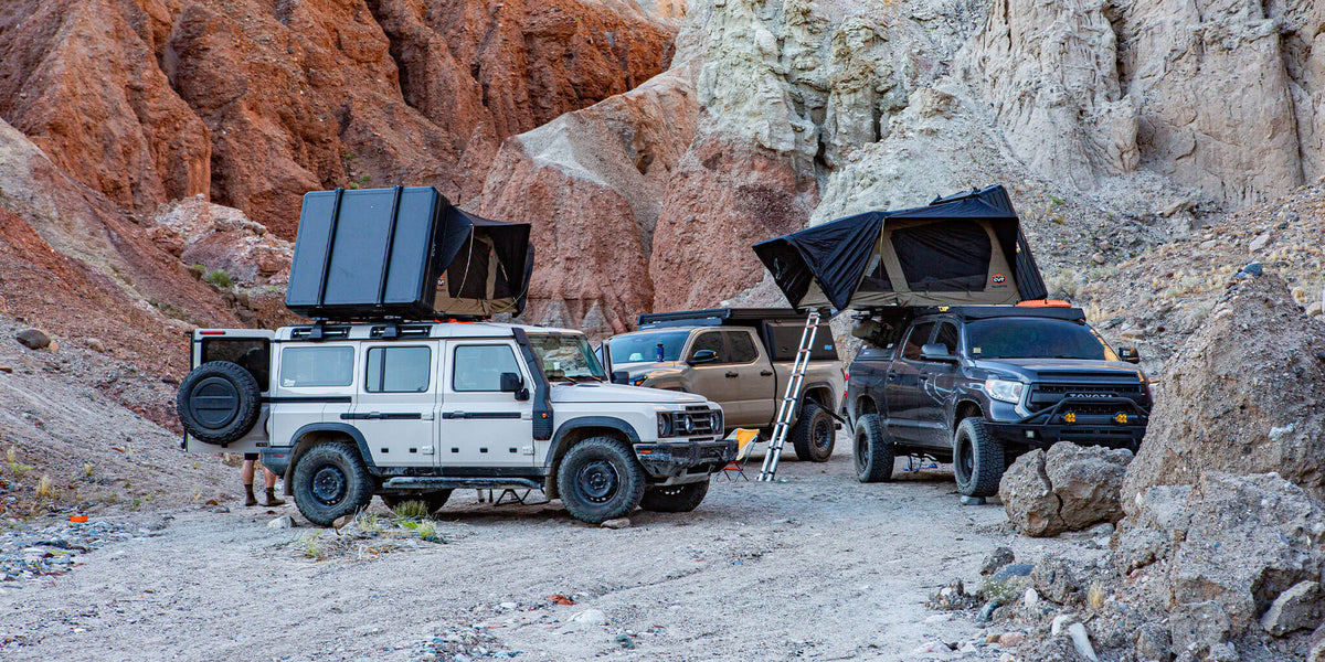 Three off-road vehicles with rooftop tents are parked in a rocky canyon. The tents are open, and a ladder leans against one vehicle. The colorful canyon walls surround the small campsite.