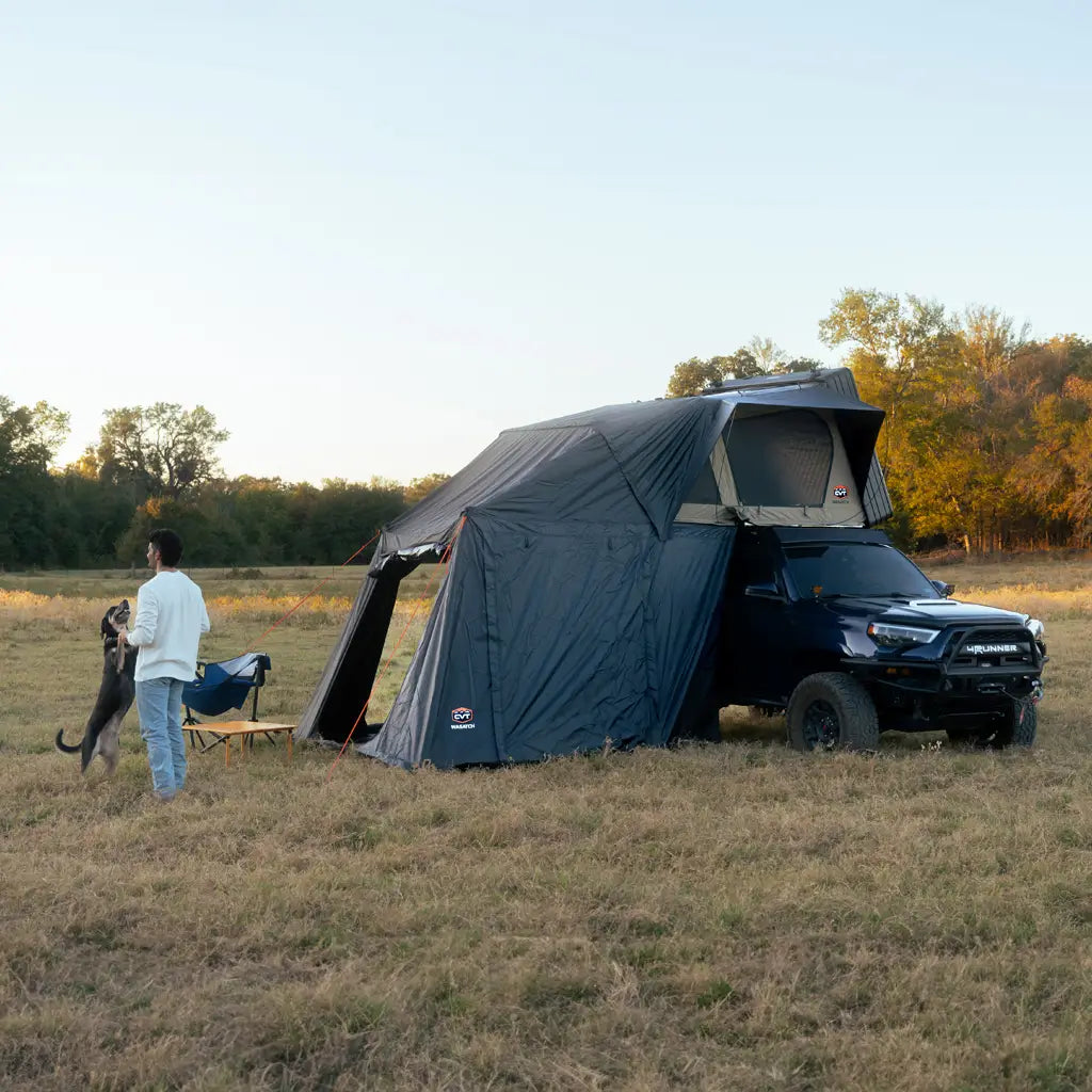 A person stands with a dog near a black SUV outfitted with a rooftop tent and awning, parked in an open grassy field at sunset.