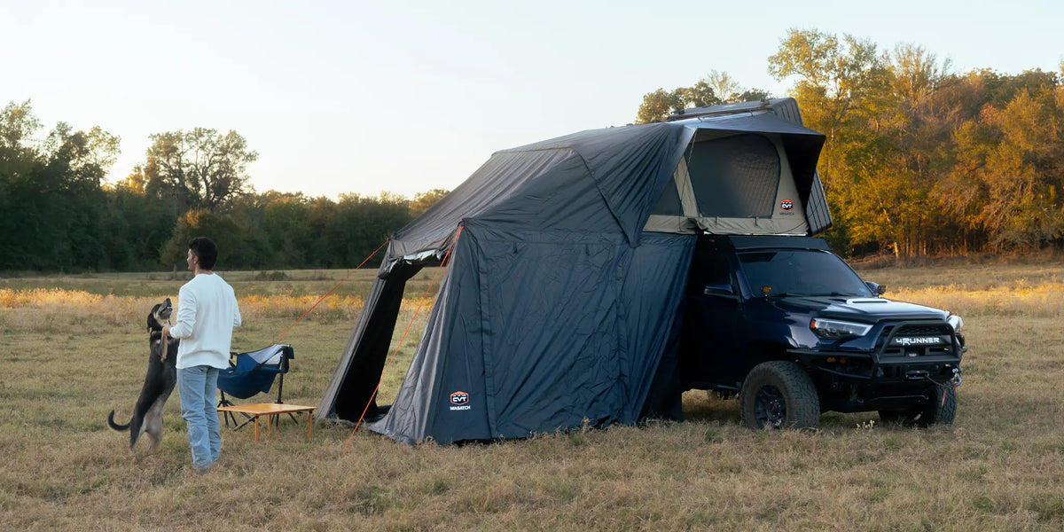 A person stands in a grassy field with a dog near a black SUV equipped with a rooftop tent and awning. Nearby are a camping chair and table. Trees and golden light are in the background.