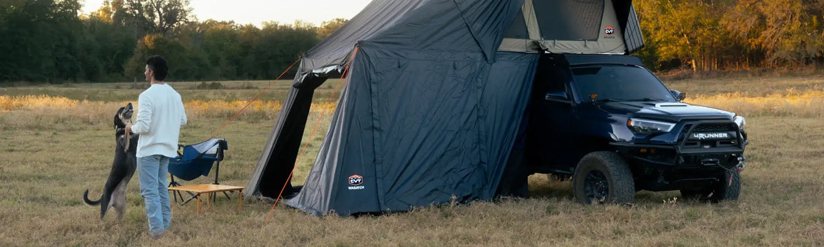 A person stands in a field playing with a dog next to a black SUV with a rooftop tent and attached annex. Nearby, a chair and a small wooden table are set up. Trees and tall grass are visible in the background.