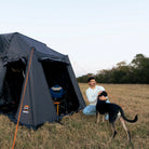 A man kneels on the grass beside a CVT Wasatch Annex shelter, petting a black and tan dog. Camping gear is set up inside the annex, providing rugged protection for outdoor adventures. Trees and open sky create a scenic backdrop.