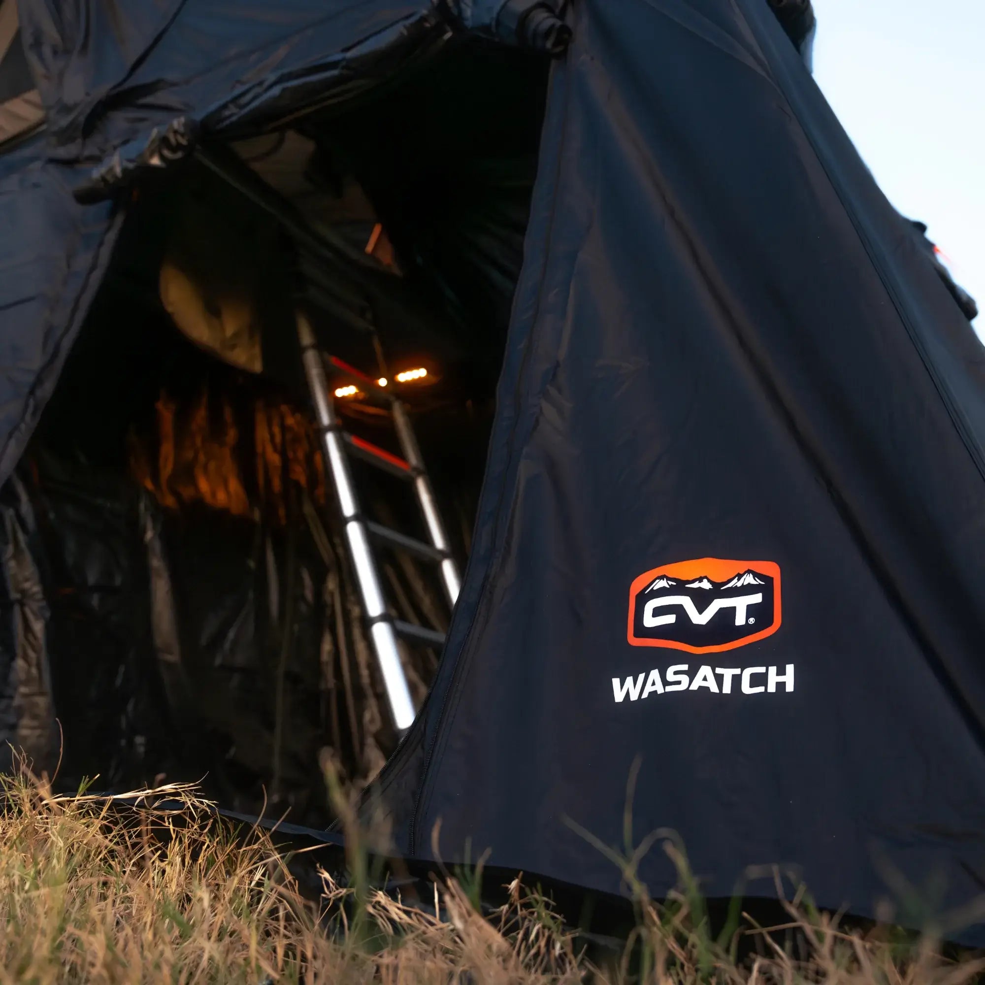 A close-up of a black tent with CVT WASATCH branding, partially open to reveal an aluminum ladder inside and interior lights glowing. Grass is visible in the foreground.