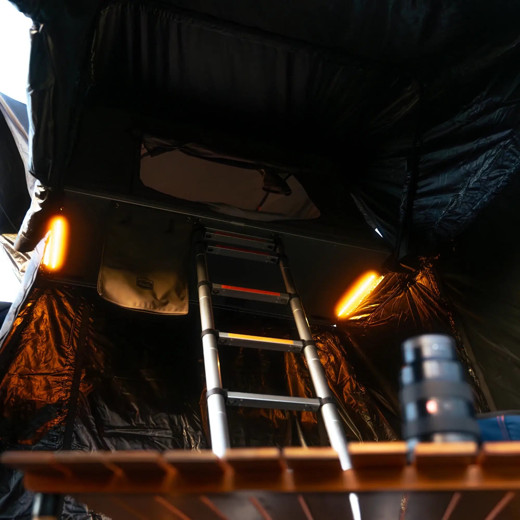 A ladder leads up to a rooftop tent with glowing orange lights inside. A wooden table and a camera lens are visible in the foreground.