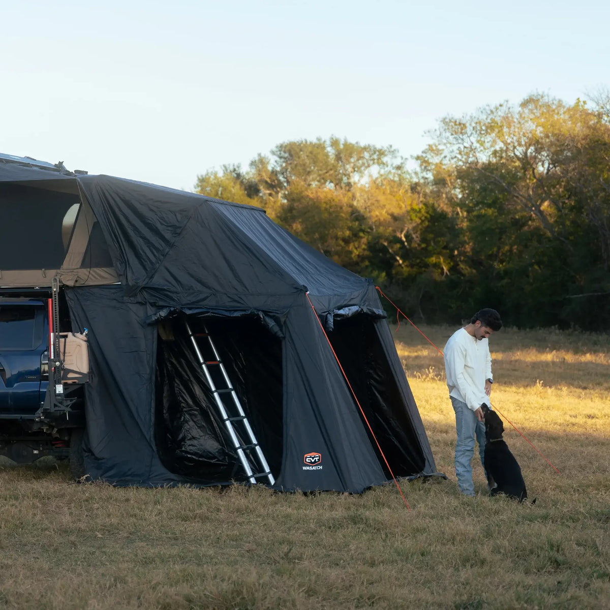 A man stands in a grassy field next to his vehicle, which is equipped with a CVT Wasatch Annex—a durable rooftop tent from CVT that provides dependable shelter for camping adventures. He is petting a black dog, while trees and a clear sky can be seen in the background.
