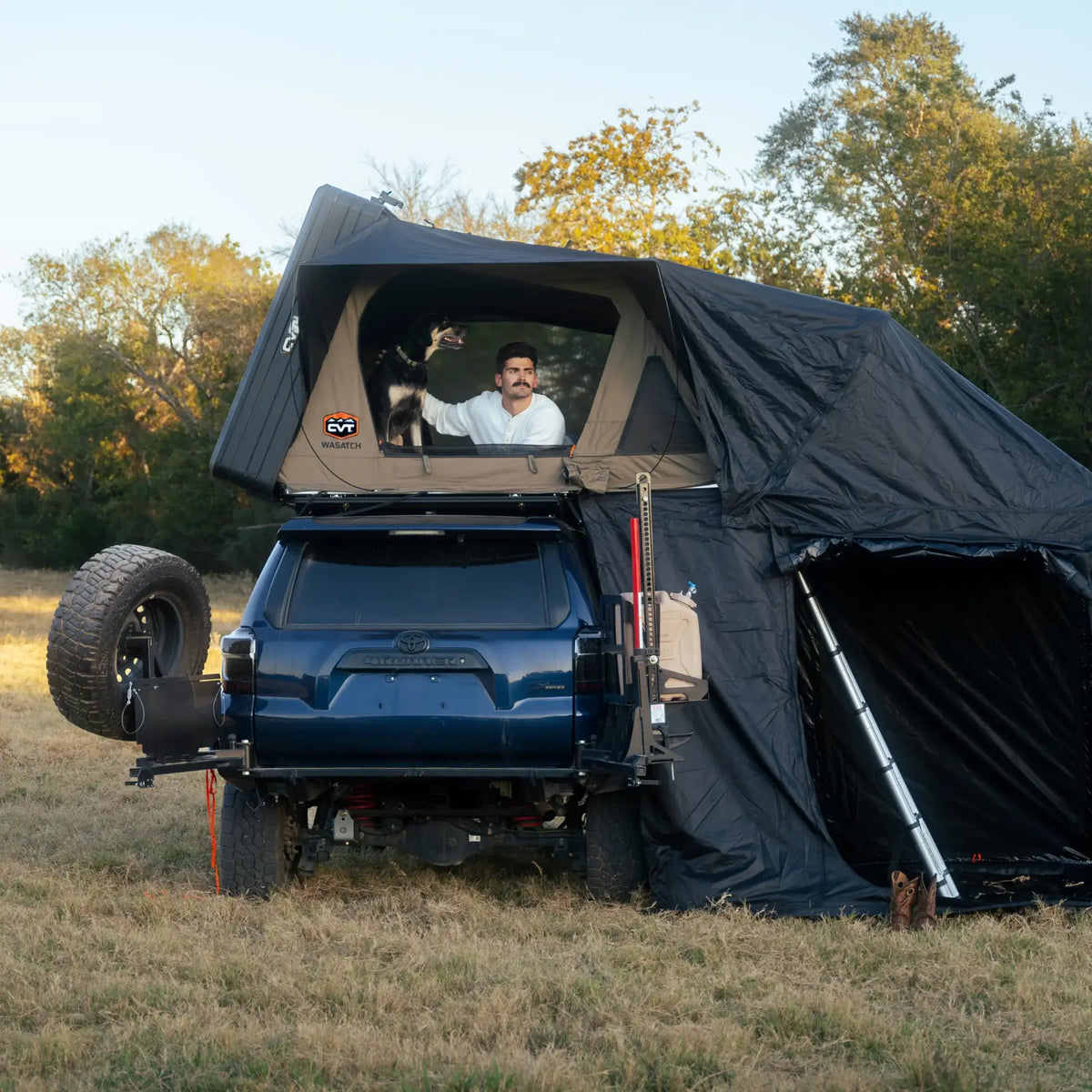 A man and a dog are in a rooftop tent mounted on a blue SUV, parked on grass with trees in the background. The SUV has camping gear and an attached ground tent.
