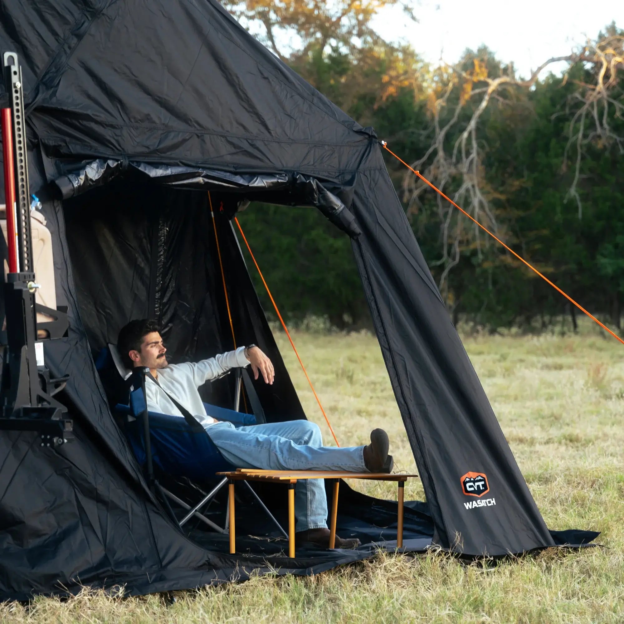 A person relaxes in a camping chair with feet up on a table inside a black tent on a grassy field, surrounded by trees.