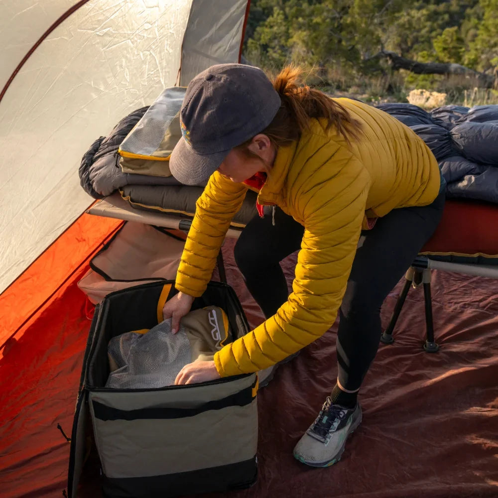 Inside a tent nestled in nature, a person in a yellow jacket and cap skillfully packs their bag using CVT Camping Cubes™, next to a cot with a sleeping bag—demonstrating how CVT’s Camping Cubes™ provide thoughtful gear organization for any outdoor adventure.