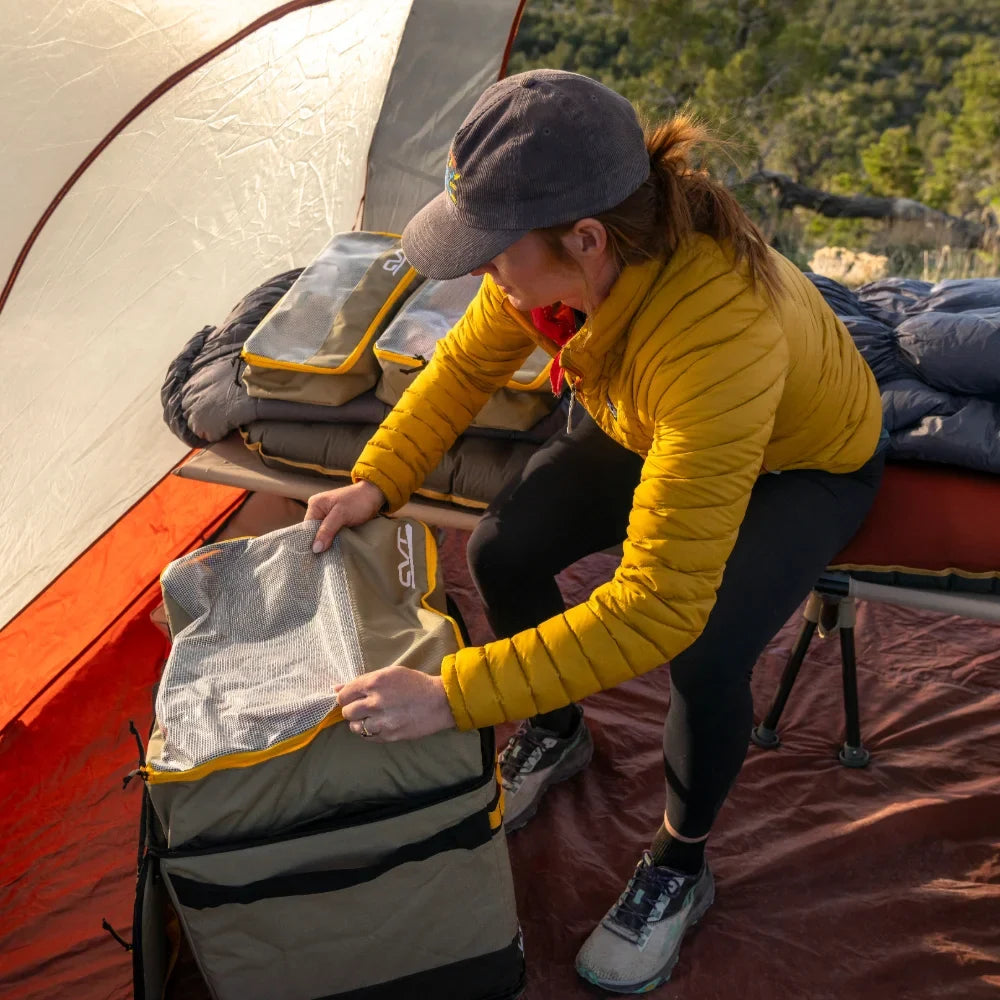 A person in a yellow jacket and cap sets up gear inside a tent, placing items in CVT Camping Cubes™ for efficient organization on a cot. Trees and nature are visible outside.