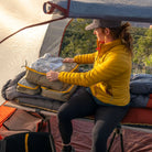 A person in a yellow jacket sits on a cot inside a tent, practicing gear organization with CVT Camping Cubes™ as they arrange bags on top of a water-resistant sleeping bag. Sunlight filters through the tent, and trees are visible outside.