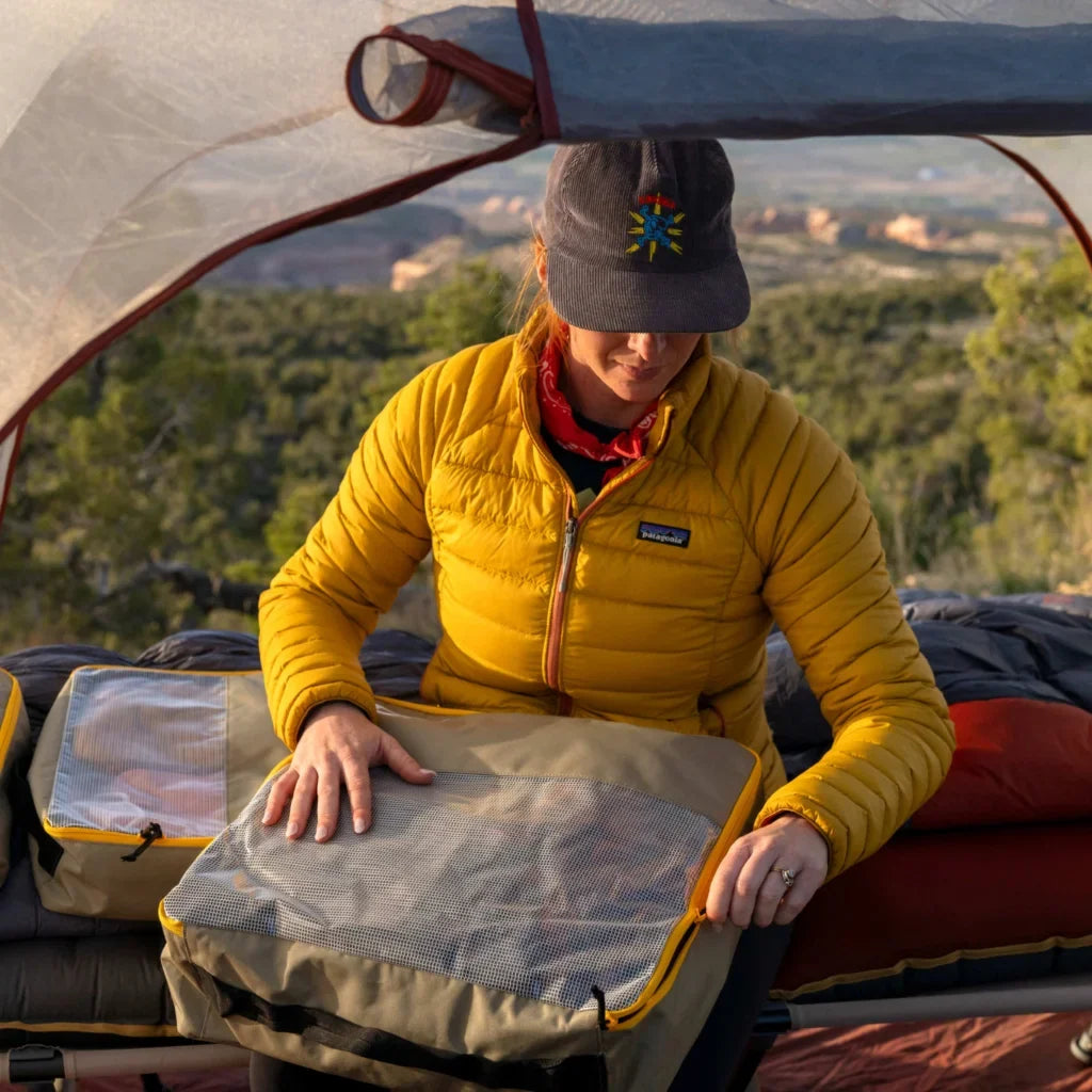 A person in a yellow jacket and cap packs gear inside a tent with a scenic, mountainous landscape and greenery visible in the background.