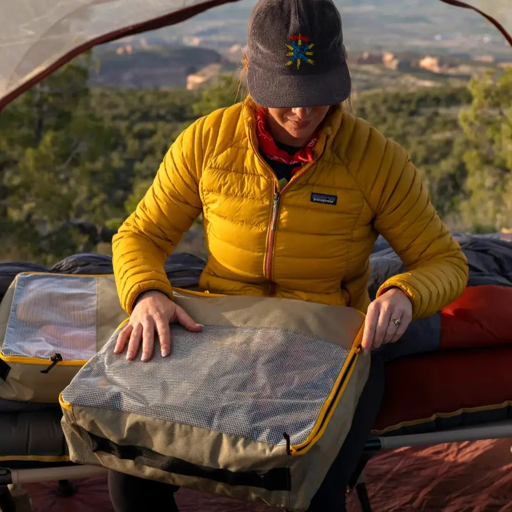 A person in a yellow jacket and cap sits in a tent, unzipping a CVT Camping Cubes™ storage cube on a cot, with trees and mountains visible in the background.