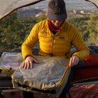 A person in a yellow jacket and cap sits in a tent, unzipping a CVT Camping Cubes™ storage cube on a cot, with trees and mountains visible in the background.