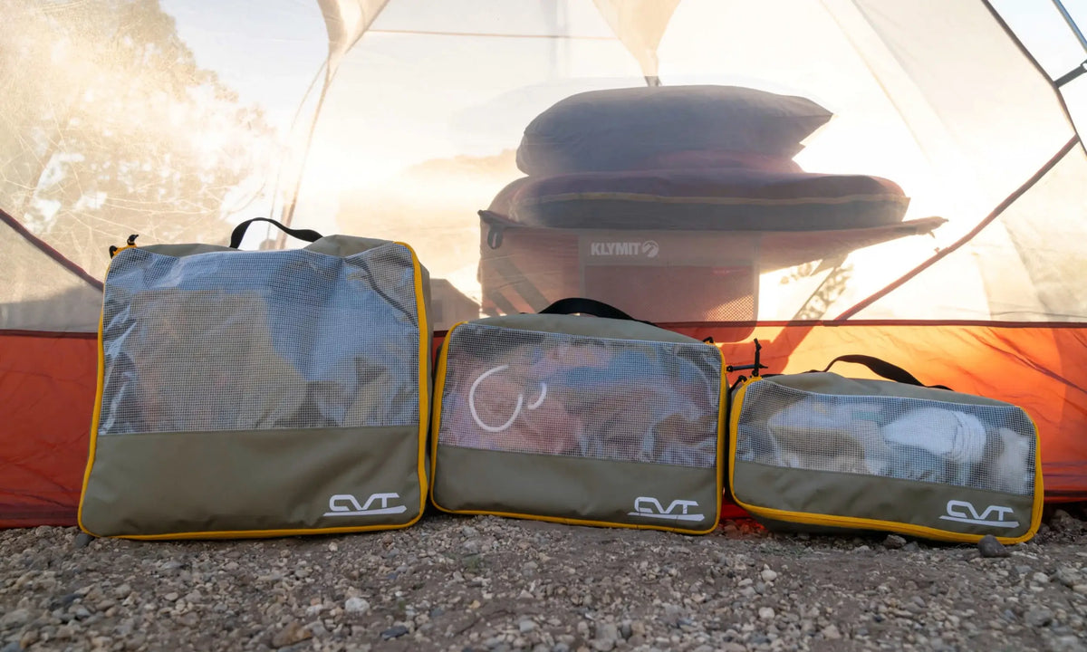 Three packed mesh travel bags sit on rocky ground inside a tent, with sleeping pads and pillows visible in the background. Sunlight filters through the tent fabric.