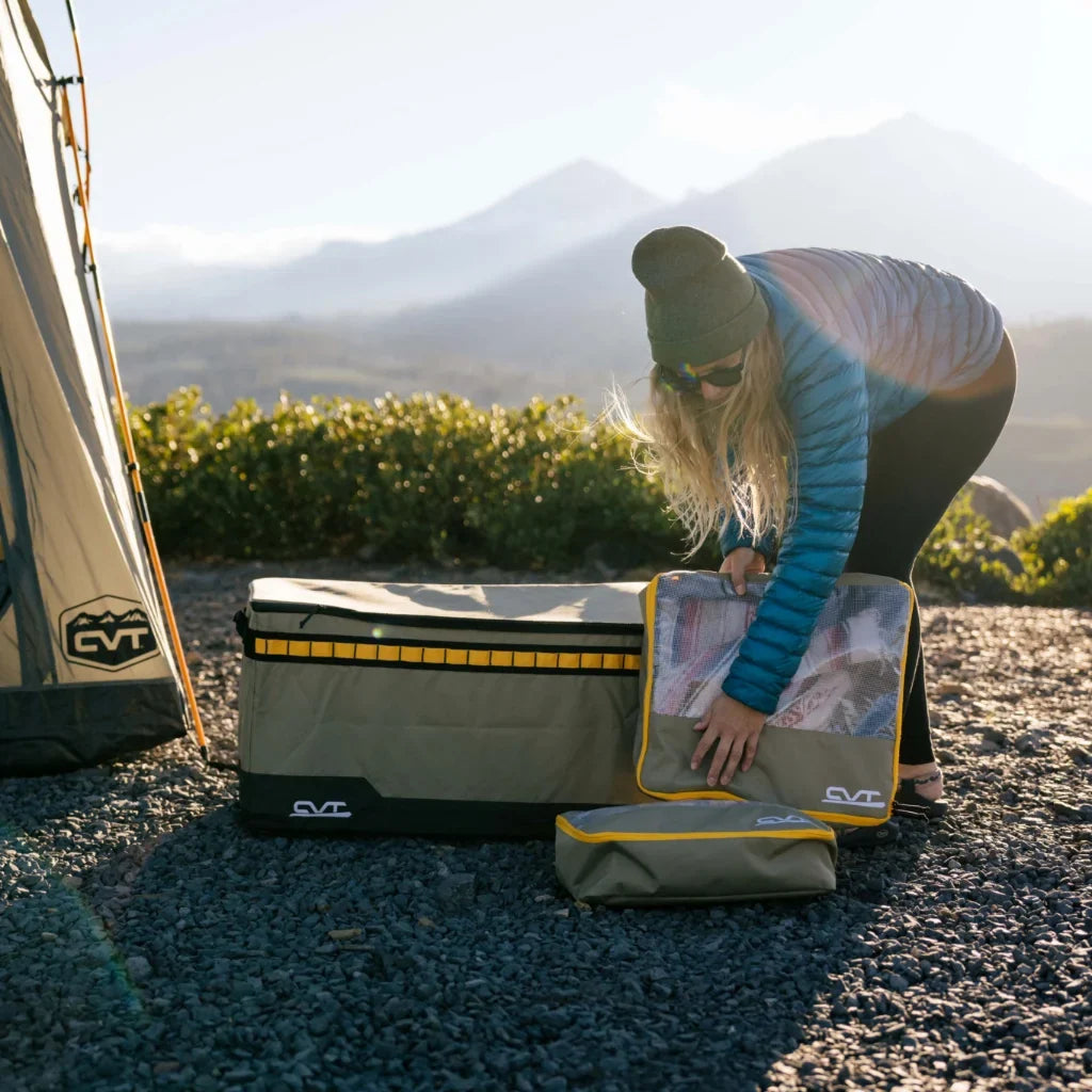 A person wearing sunglasses and a beanie organizes camping gear in storage bags next to a tent, with mountains and sunlight in the background.
