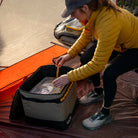 Outfitted in outdoor gear, a person sits in a tent and opens the CVT Camping Cubes™—a water-resistant, insulated cooler bag with yellow trim—next to a sleeping bag on the ground, demonstrating smart organization for an efficient camping setup.