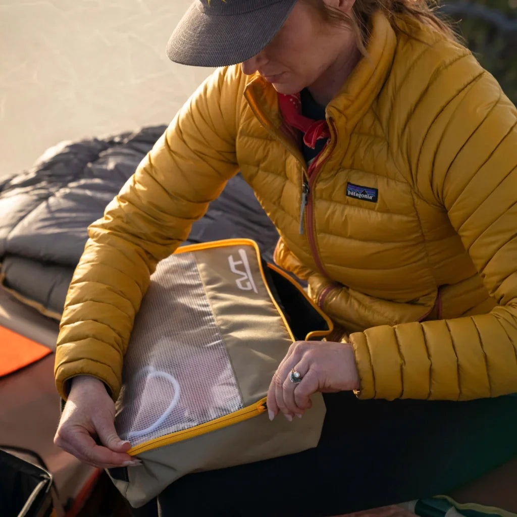 A person in a yellow jacket sits by a sleeping bag and unzips a beige and mesh packing cube, possibly while camping outdoors.