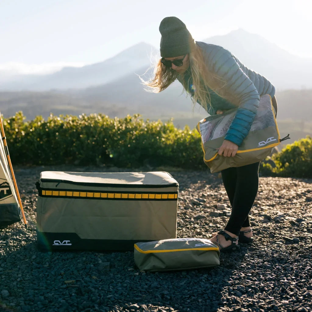 A person in outdoor gear sets up camping storage bags on rocky ground with mountains and greenery in the background, under soft sunlight.