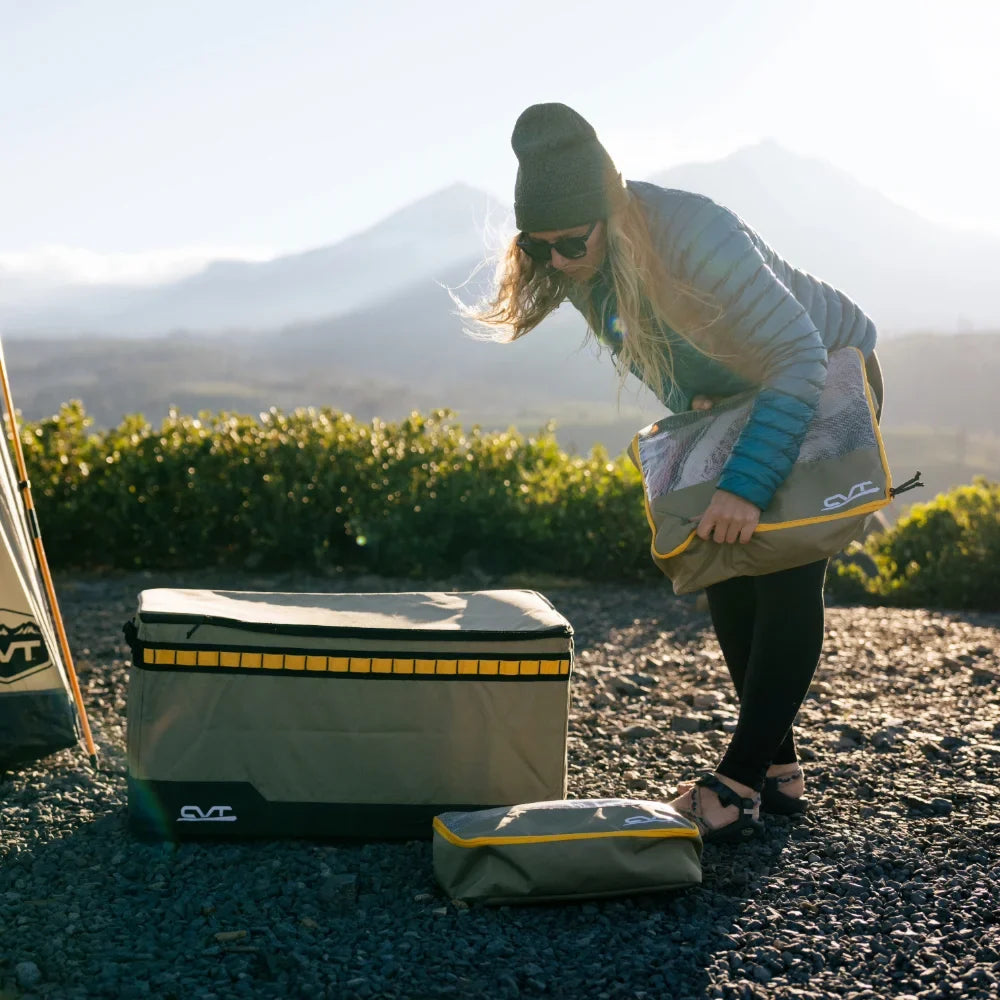 Clad in outdoor clothing and a beanie, an individual unpacks CVT Camping Cubes™—water-resistant organizers designed for efficient gear storage—on rocky terrain, with scenic mountains and lush greenery behind them.