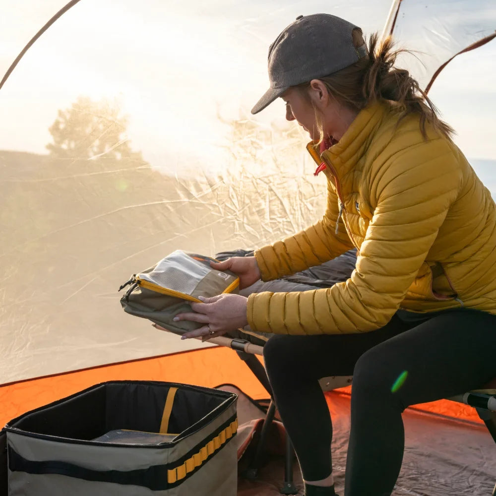 A person wearing a yellow jacket and cap sits inside a tent, organizing gear from a storage bin with the help of CVT Camping Cubes™, while morning sunlight filters through the water-resistant tent fabric.