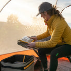 A person wearing a yellow jacket and cap sits inside a tent, organizing gear from a storage bin with the help of CVT Camping Cubes™, while morning sunlight filters through the water-resistant tent fabric.