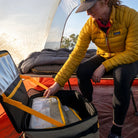 A person in a yellow jacket kneels inside a tent, reaching into a CVT Camping Cubes™ soft-sided cooler designed for gear organization. Behind them are a cot and sleeping bag, with sunlight filtering through the water-resistant tent walls.
