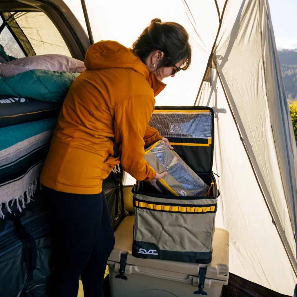 A person in an orange jacket and sunglasses places a silver bag into a gray and yellow cooler inside a tent filled with blankets and camping gear.