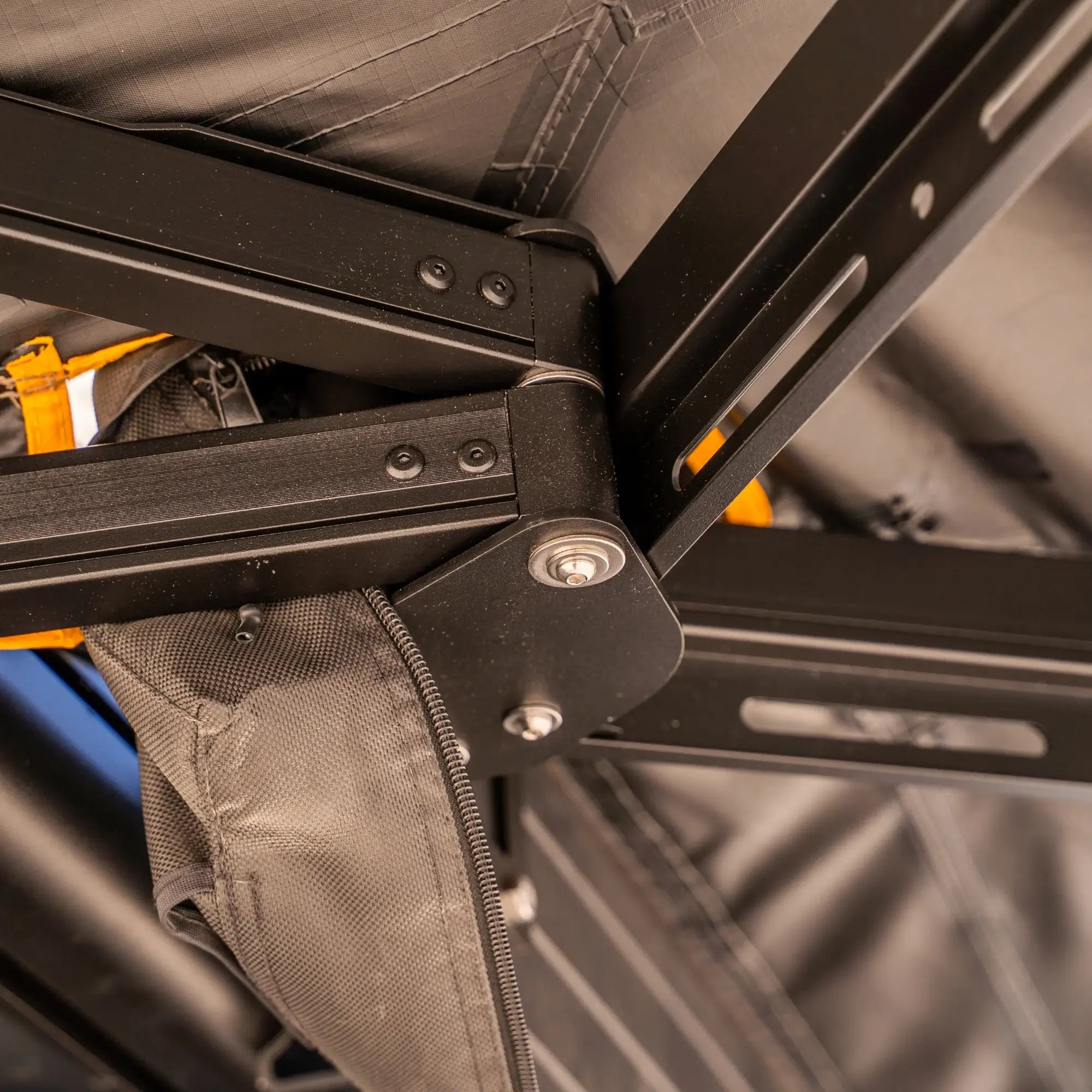 Close-up of a black metal frame structure with bolts, supporting a gray fabric canopy. Part of an orange strap and a zippered fabric compartment are also visible.