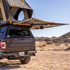 A pickup truck equipped with a rooftop tent and the CVT 270 Degree Awning, featuring a batwing design, is parked in a dry, rocky desert landscape under a clear blue sky.