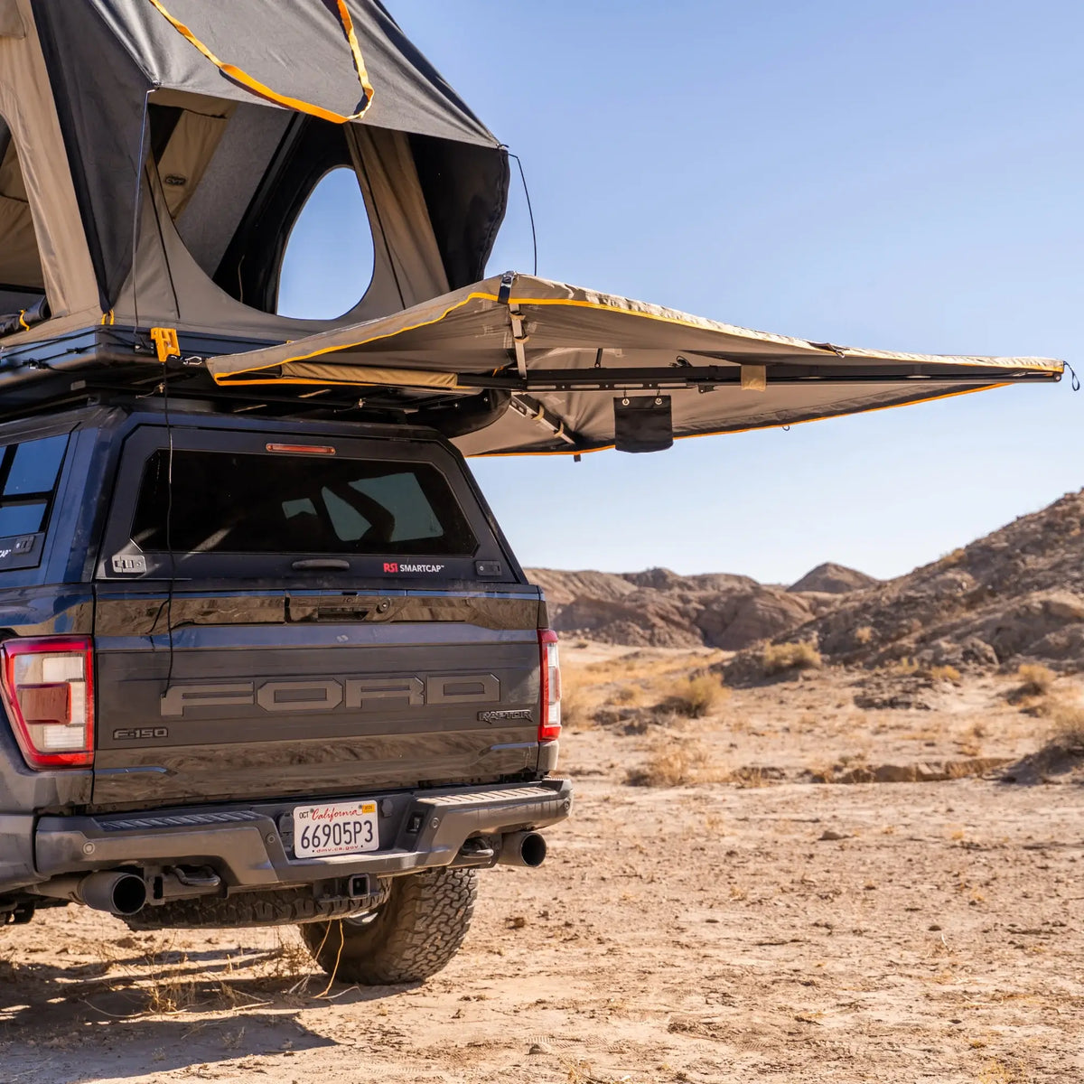 A pickup truck equipped with a rooftop tent and the CVT 270 Degree Awning, featuring a batwing design, is parked in a dry, rocky desert landscape under a clear blue sky.