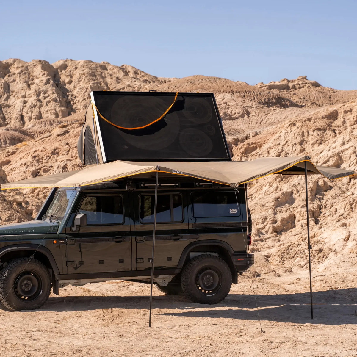 A dark green SUV equipped with a CVT 180 Degree Awning and a rooftop tent is parked in a rocky desert area under a clear sky, providing instant shade and outdoor shelter for any adventure.