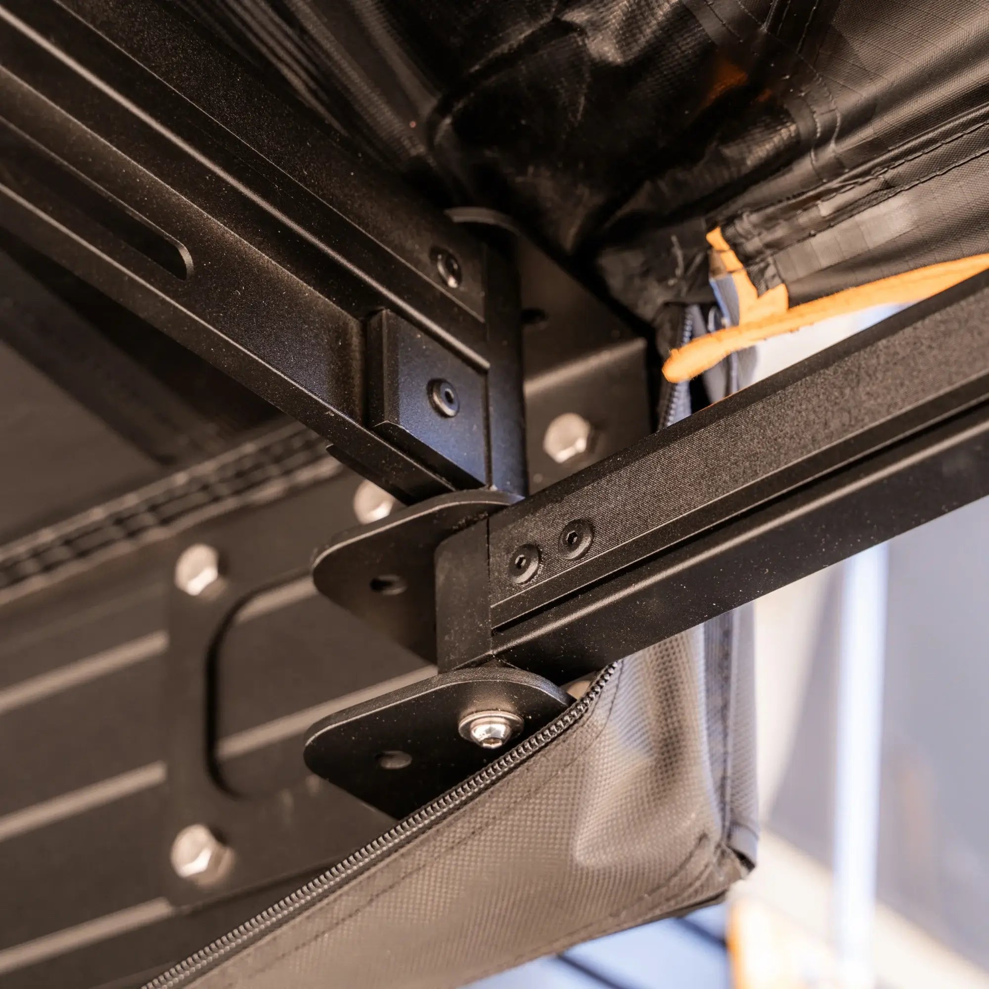 Close-up of black metal bracket hardware connecting an awning frame to a roof rack, with visible bolts, screws, and canvas fabric with an orange zipper pull.