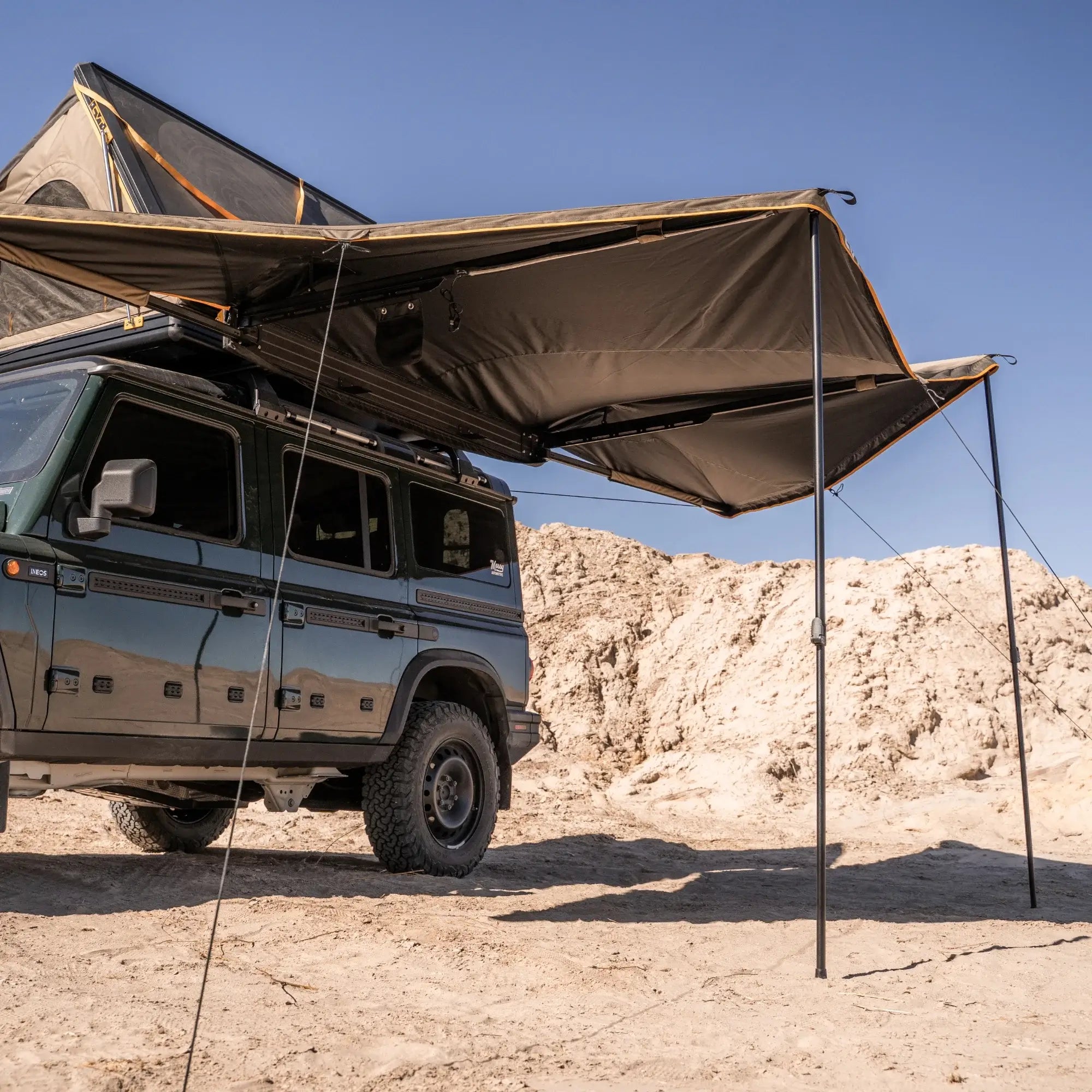 A rugged off-road vehicle with a rooftop tent and large awning is parked on sandy terrain under a clear blue sky, with rocky hills in the background.