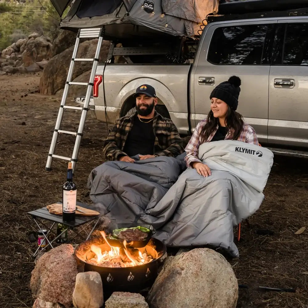 Two people relax by a campfire, wrapped in cozy gray sleeping bags and the Klymit Horizon™ Overland Blanket from Klymit—a water resistant layer of comfort—seated in front of a truck outfitted with a rooftop tent. A small table with wine and food completes their enjoyable outdoor camping scene.