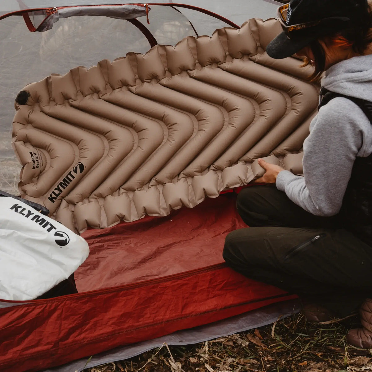 A person kneels beside a red tent, holding an inflated tan Klymit Insulated Static V™ Sleeping Pad. A white Klymit bag is nearby, and the setting appears to be outdoors in a natural area.