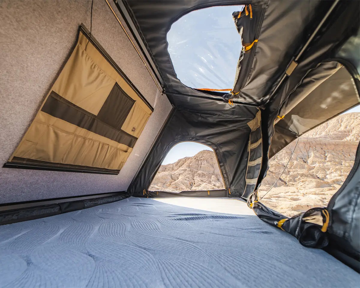 Interior view of a rooftop tent with grey walls, tan storage pockets, large windows, and a blue sleeping surface. Rocky desert landscape visible through the windows.