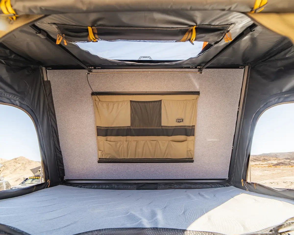 View from inside a rooftop tent, showing a flat sleeping area, a storage pocket on the back wall, and windows with sunlight and desert landscape visible outside.