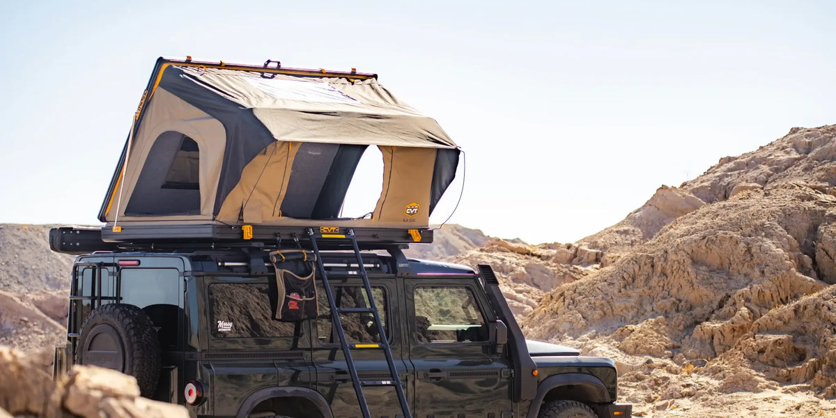 A rooftop tent is set up on top of a parked black SUV in a rocky desert landscape, with a ladder leading up to the tent.